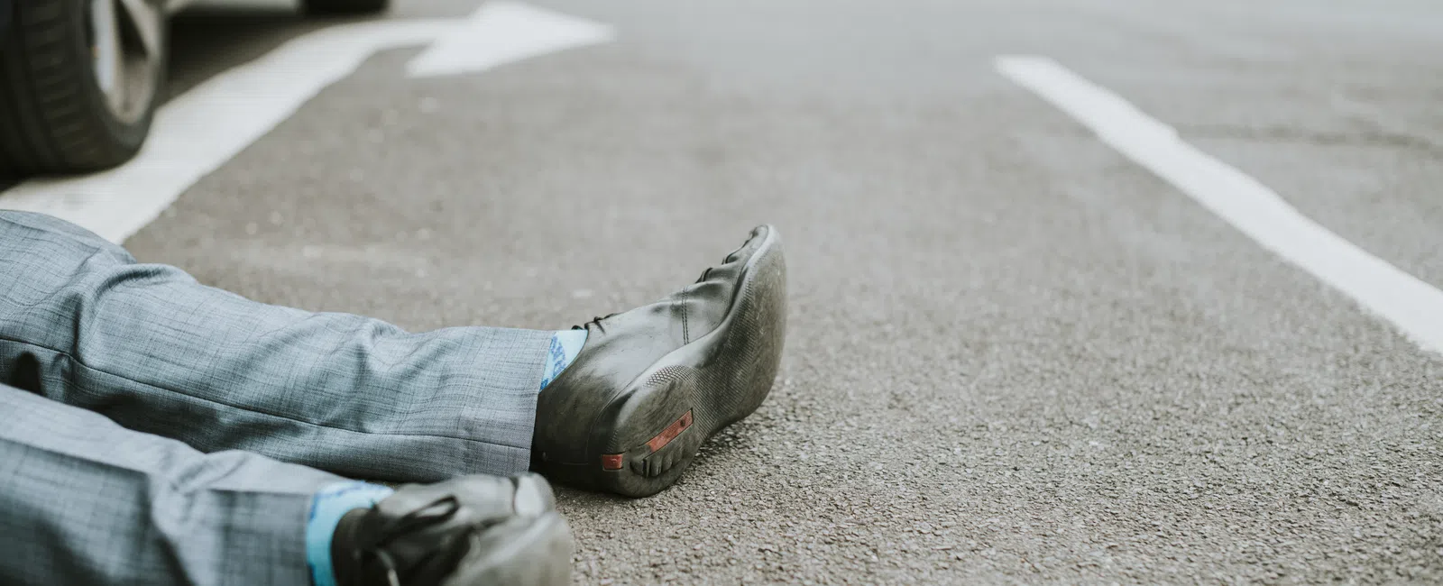 Man lying on street wearing gray suit and black shoes, viewed from waist down on asphalt road.