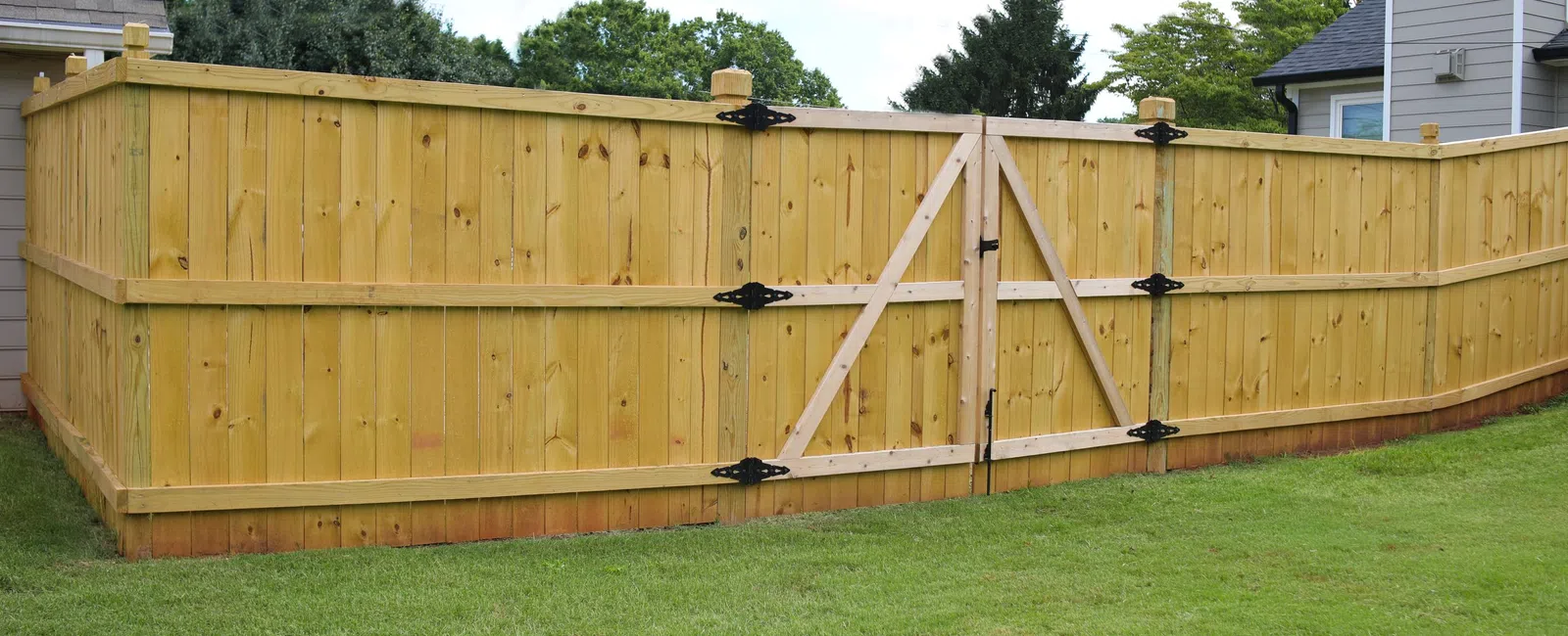 New wooden privacy fence with double gate installed around a backyard lawn area between houses.
