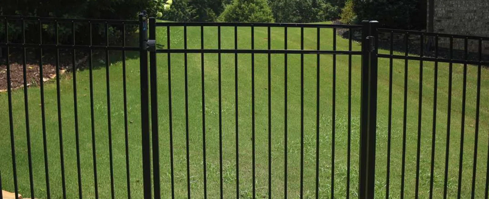 Black metal fence gate in front of green lawn with bushes and trees under a blue sky.