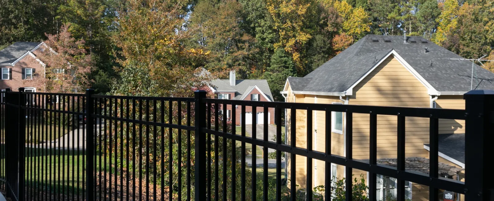 Black metal fence overlooking homes surrounded by colorful autumn trees under a clear blue sky.