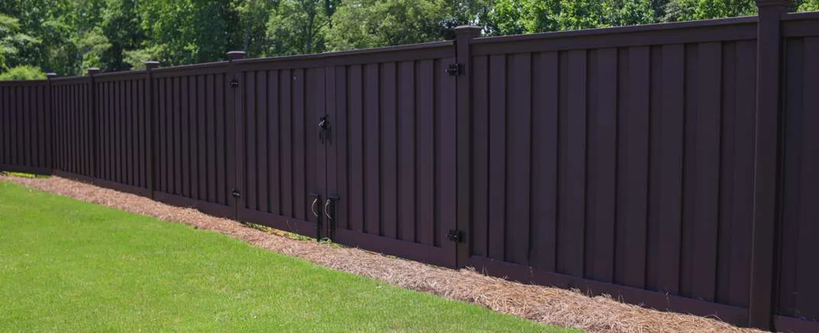 Long dark brown wooden fence with gate enclosing a grassy yard next to green trees in sunlight
