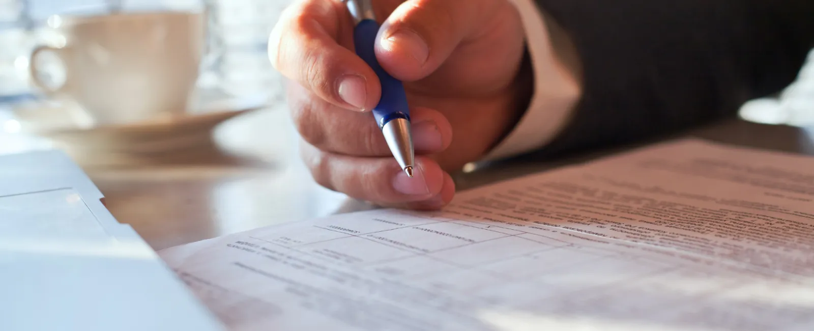 A close-up of a person signing paperwork at a desk, with a cup of coffee in the background.