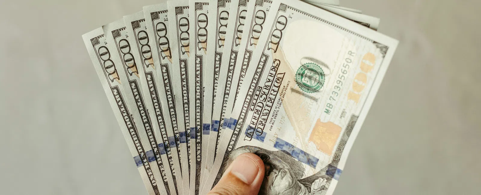 Hand holding a fan of multiple US hundred-dollar bills with a plain blurred background.