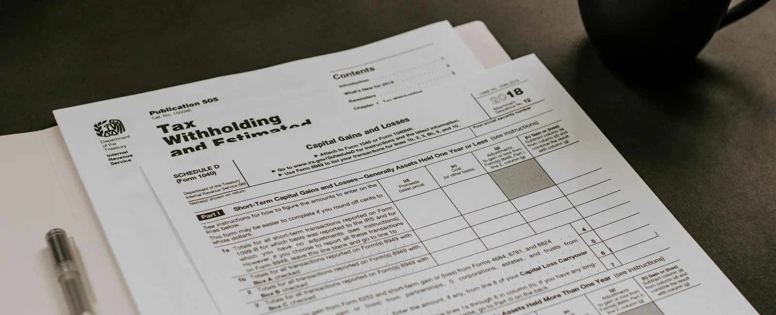 Tax withholding and estimated tax forms with pen on folder and coffee cup on dark surface.