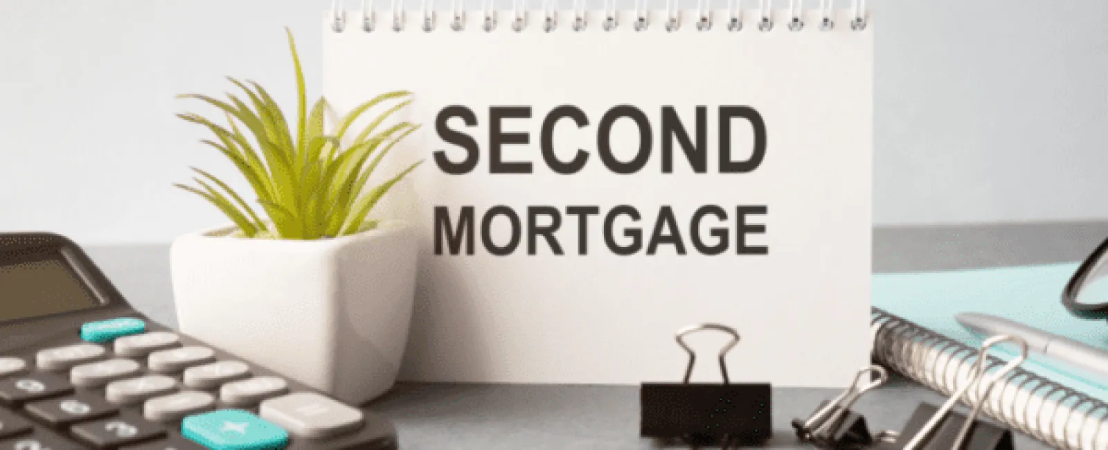 Desk setup with a calculator, small plant, binder clips, notebook, and a sign reading SECOND MORTGAGE.