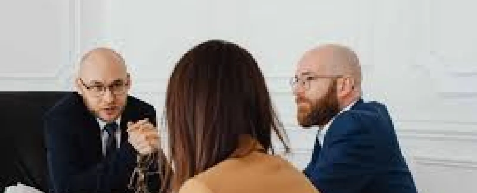 Two men in suits consulting a woman in a brown jacket at an office desk with legal books and scales of justice.