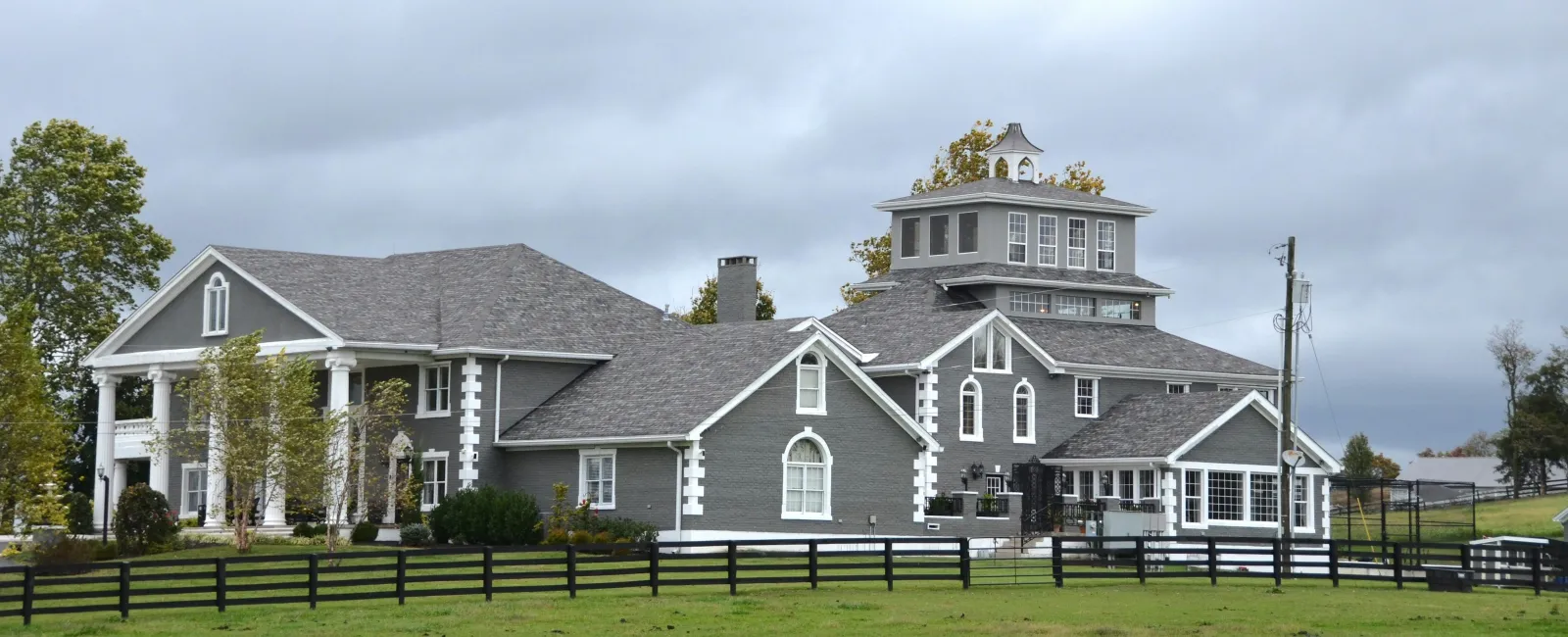 Large gray country estate with multiple peaked roofs surrounded by black fence and green lawn under cloudy sky