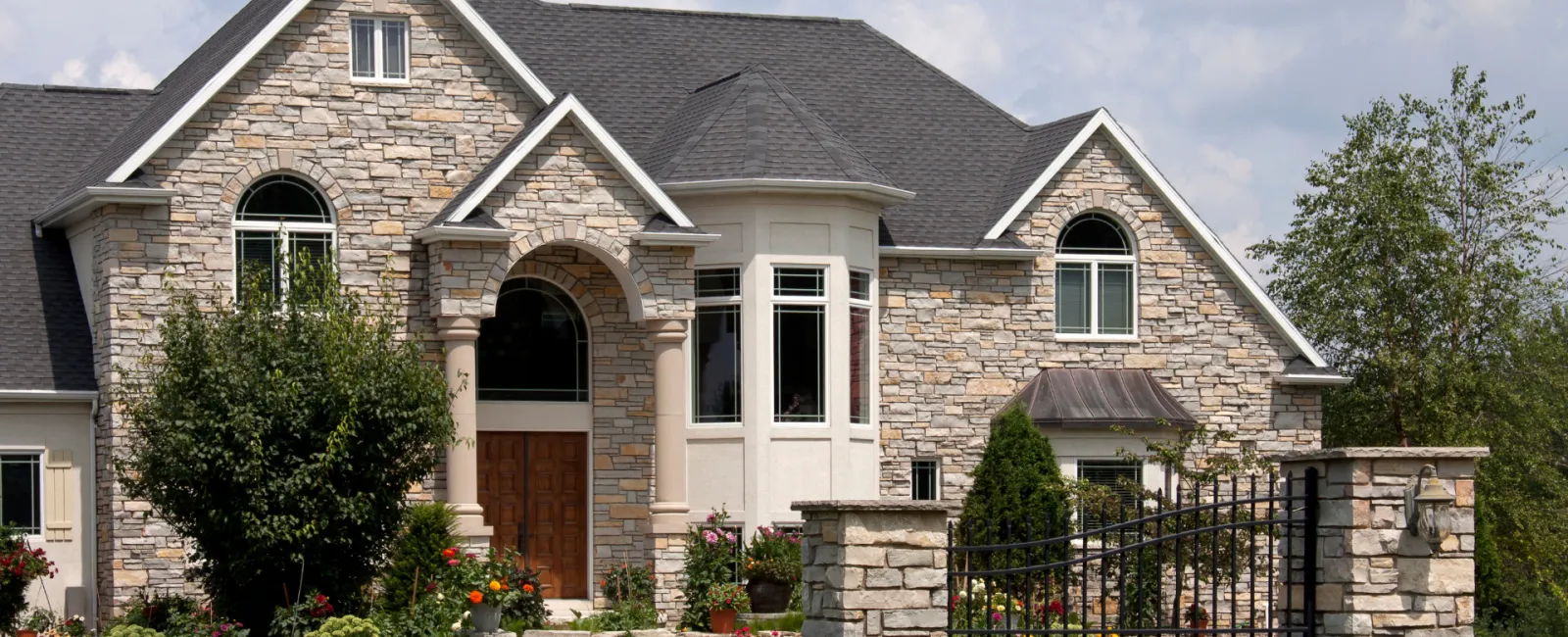 Elegant stone house with a landscaped garden and decorative gate under a bright sky.