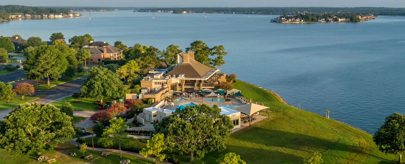 Aerial view of a lakeside resort with a pool, green lawns, trees, and nearby islands under a clear sky.
