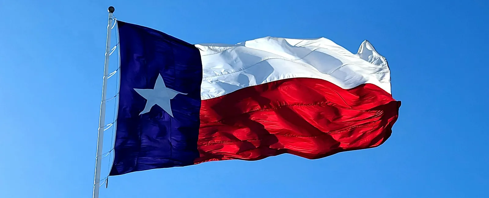 Texas state flag waving against a clear blue sky with bright sunlight enhancing its red, white, and blue colors