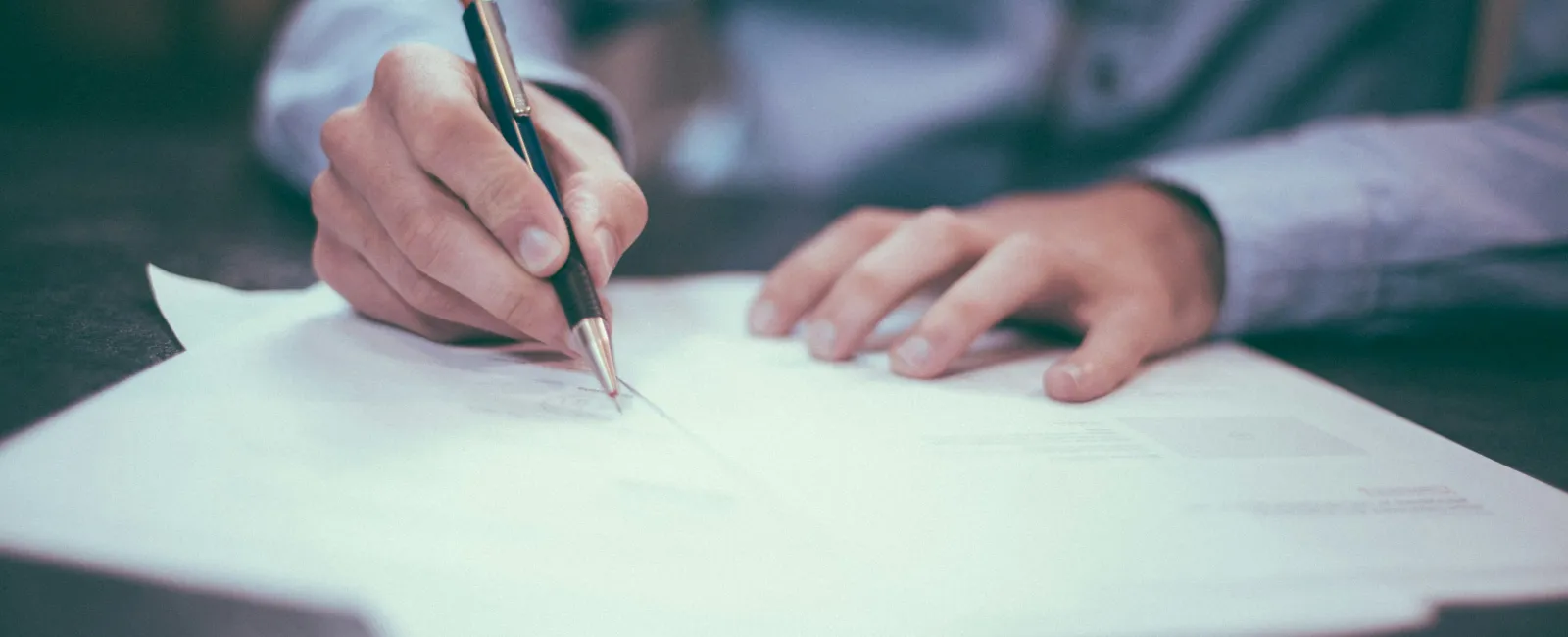 Person writing on multiple sheets of paper with a pen on a desk in soft focus background.