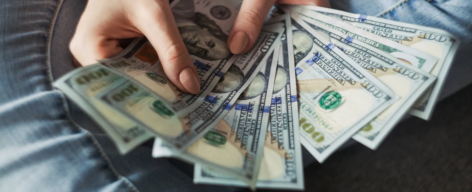 Person holding a fan of US hundred dollar bills while sitting cross-legged wearing jeans.