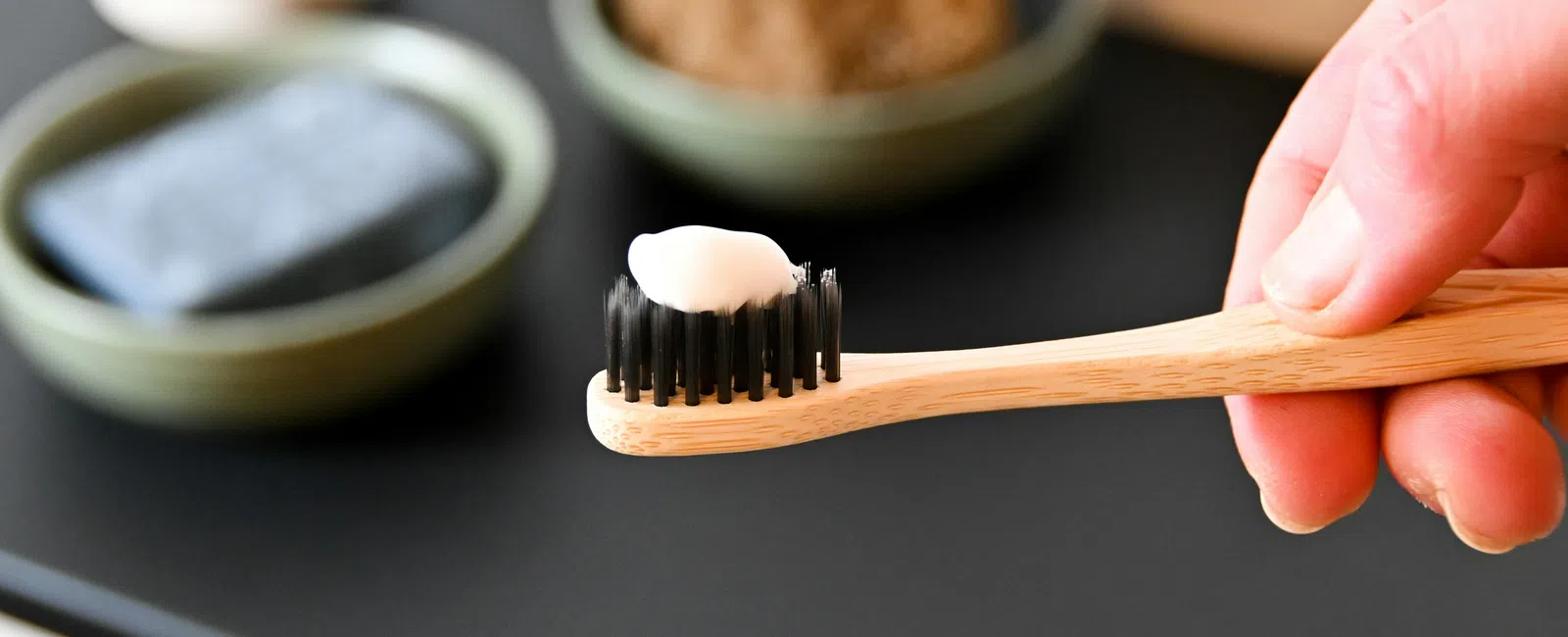 Hand holding wooden toothbrush with black bristles and white toothpaste, bathroom background with soap and sponge.