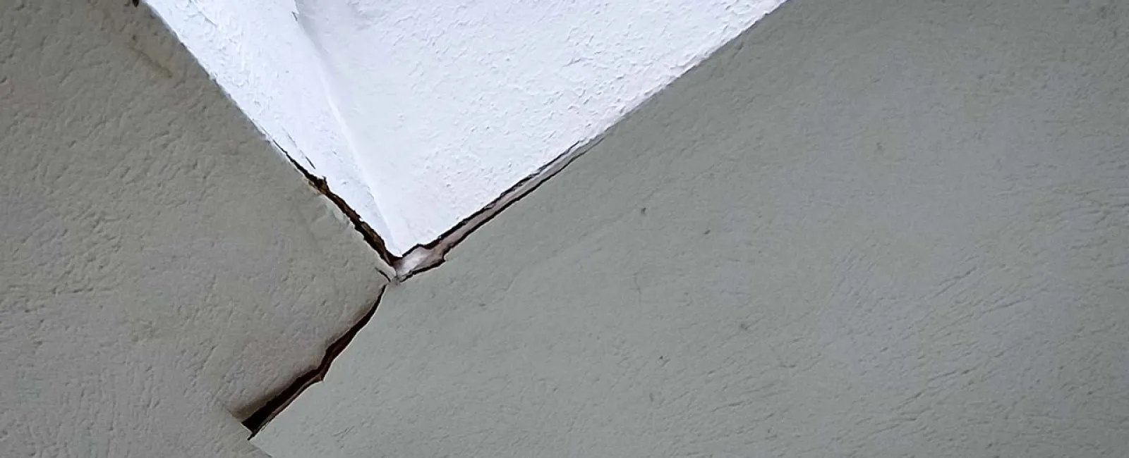 Close-up of a textured wall corner with visible cracks and paint peeling at the joint between white and beige surfaces.