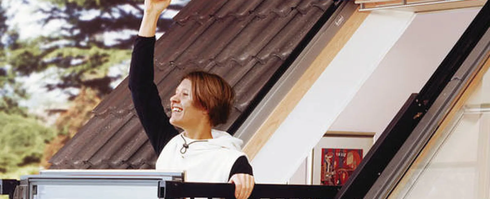 Woman standing on rooftop balcony with glass railing opening a large window on a sloped roof under clear sky