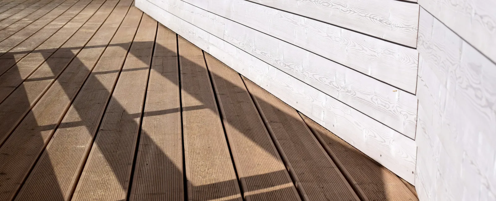 Sunlight casts geometric shadows on brown wooden deck with white horizontal siding wall.