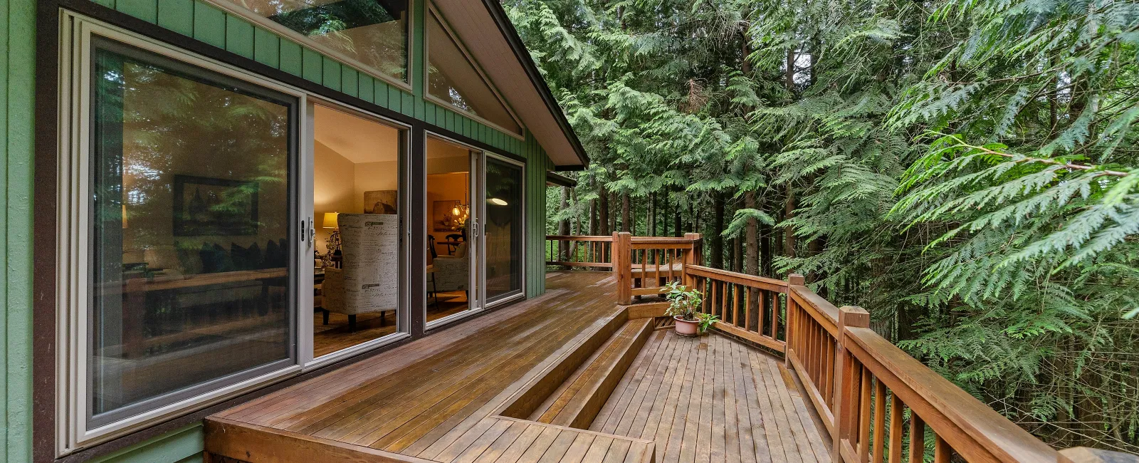 Wooden deck with built-in seating attached to green house surrounded by tall trees and plants.