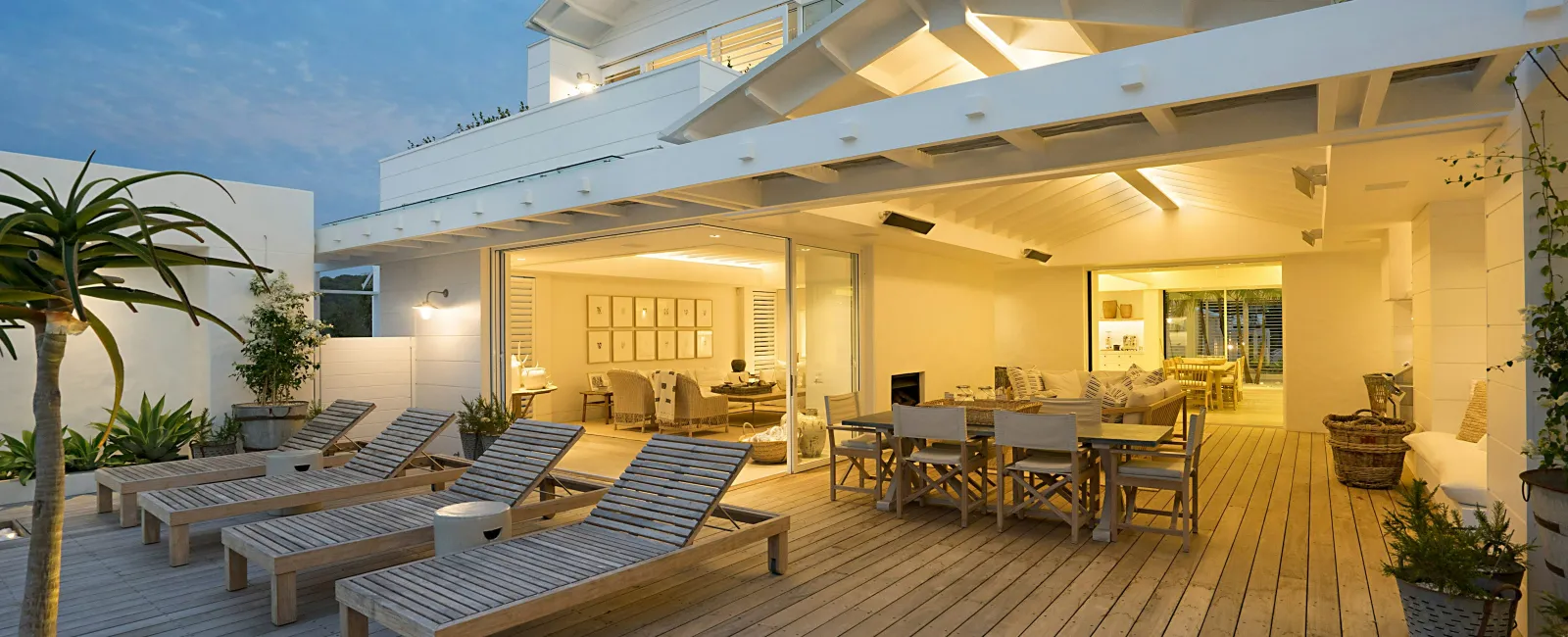 Modern outdoor patio with wooden lounge chairs, dining table, and warm indoor lighting at dusk.