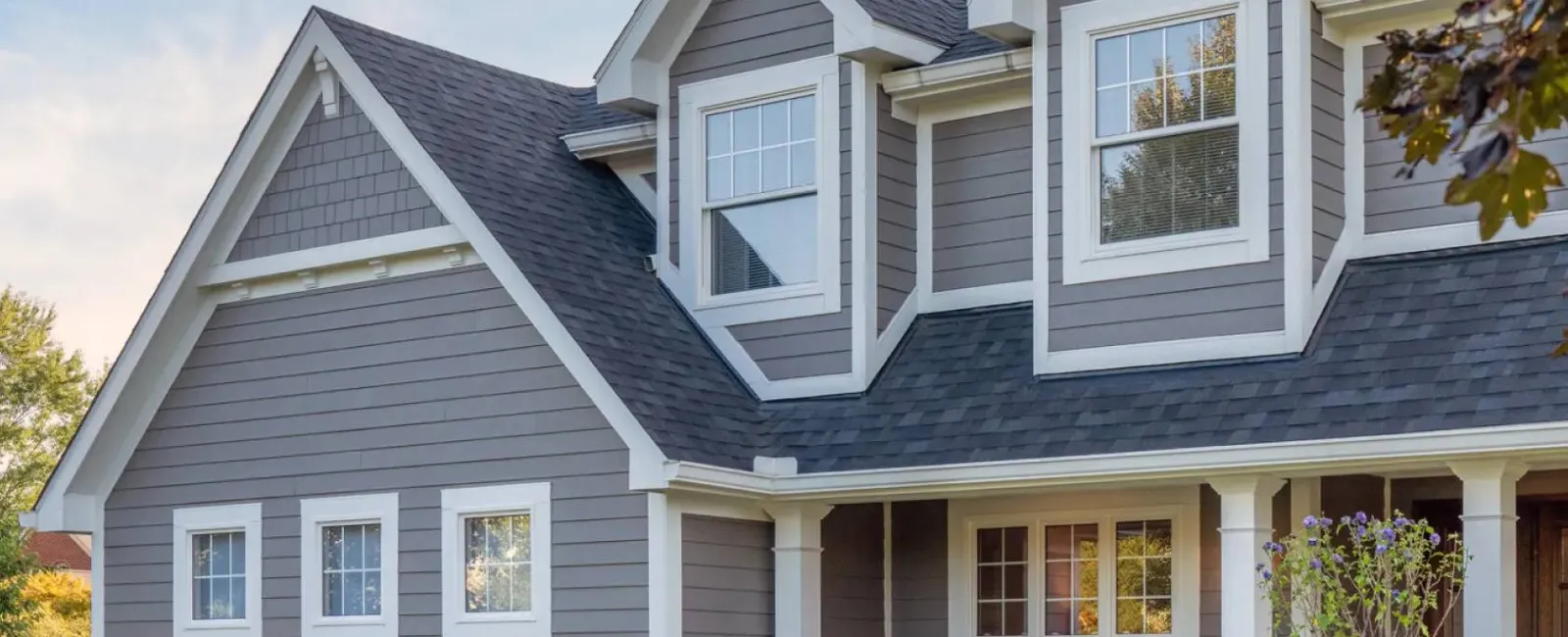 Modern gray two-story house with white trim, multiple windows, and manicured front yard with shrubs and a tree