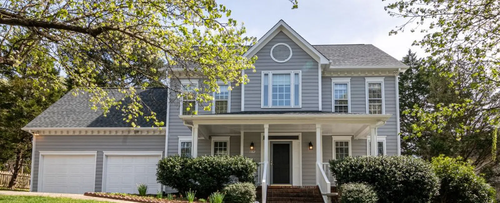 Two-story gray house with white trim, a covered front porch, and attached three-car garage surrounded by trees and green lawn.