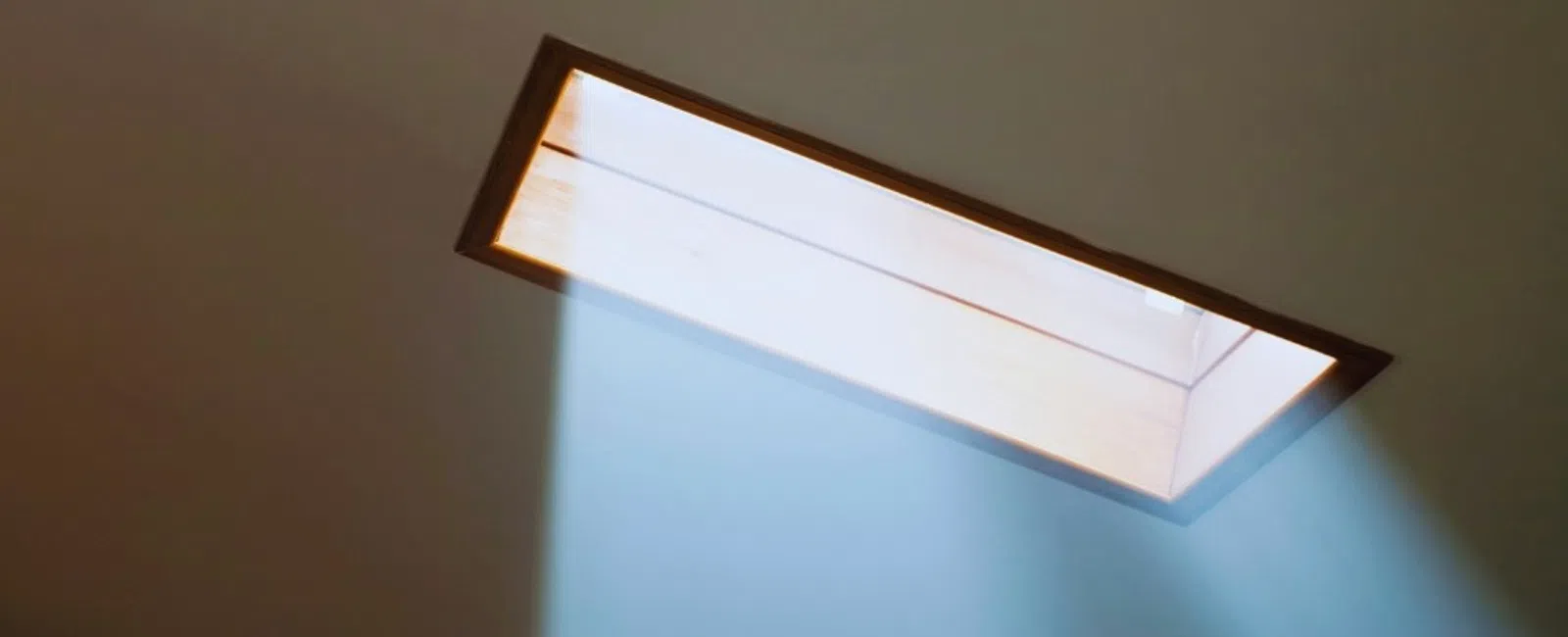 Bright sunlight beams through a rectangular skylight onto a plain ceiling casting soft light rays.