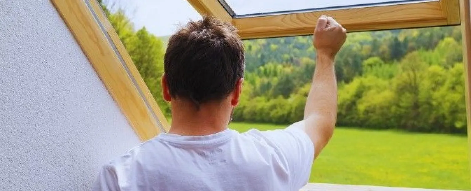 Man in white t-shirt opens a wooden framed skylight window with a scenic green landscape outside.