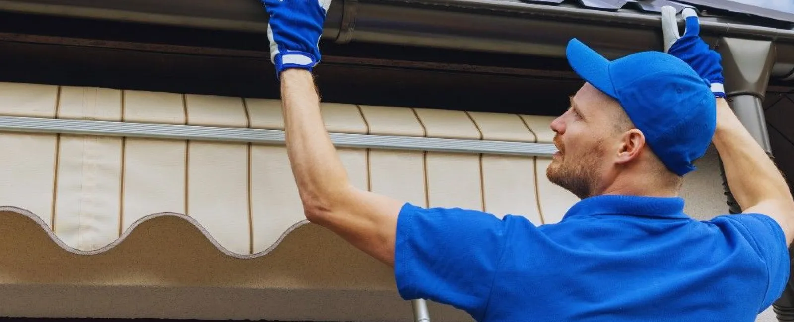 Professional worker in blue uniform installing or cleaning a metal roof gutter on a sunny day.