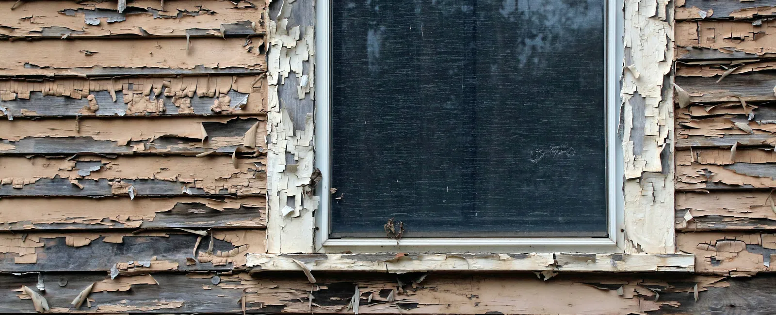 Old wooden house wall with peeling paint and a single dirty window reflecting trees and sky.