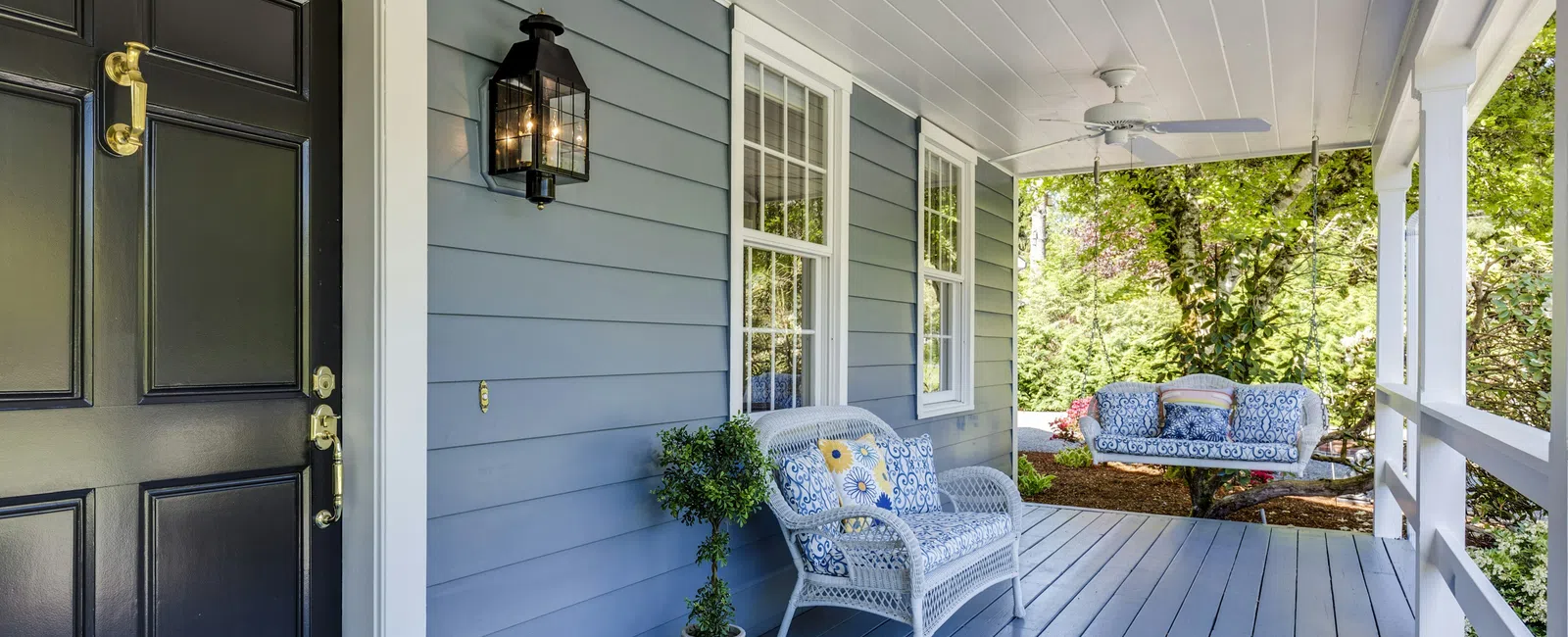 Cozy front porch with blue floor, white wicker chairs, floral cushions, black door, and surrounding greenery.