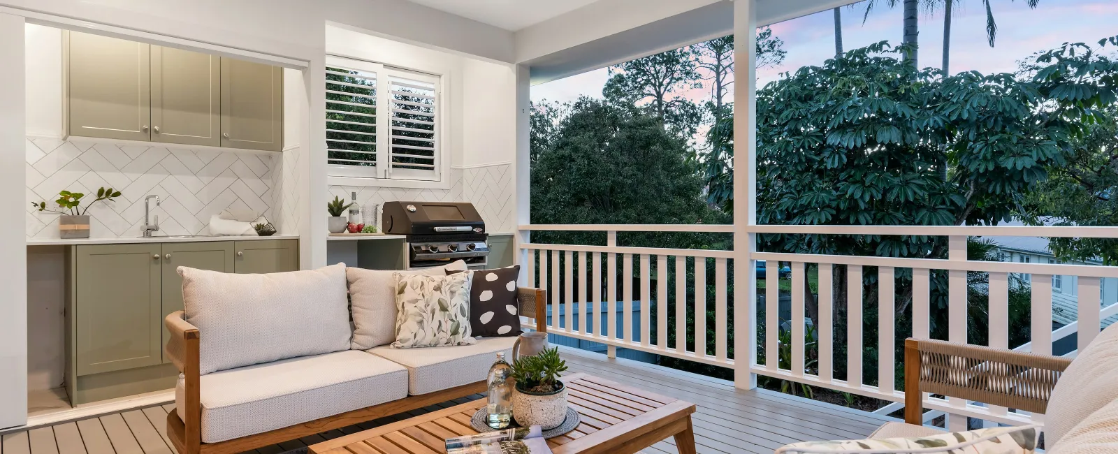Cozy outdoor patio with wooden furniture, cushions, coffee table, kitchenette, and lush greenery at sunset.