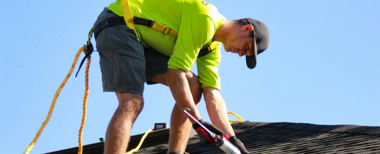 Construction worker in safety harness applying sealant on roof shingles under clear blue sky