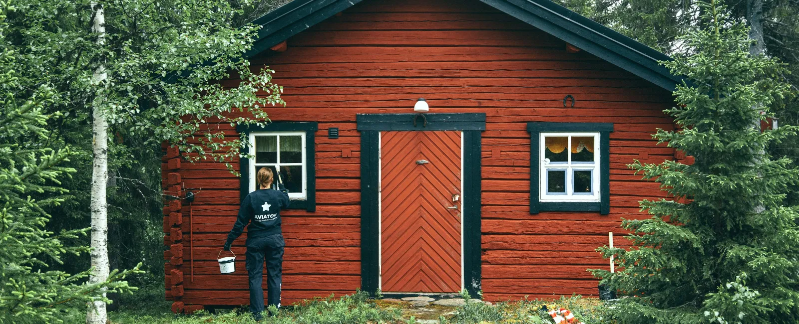 Person painting window frame of a red wooden cabin surrounded by green forest during daytime.