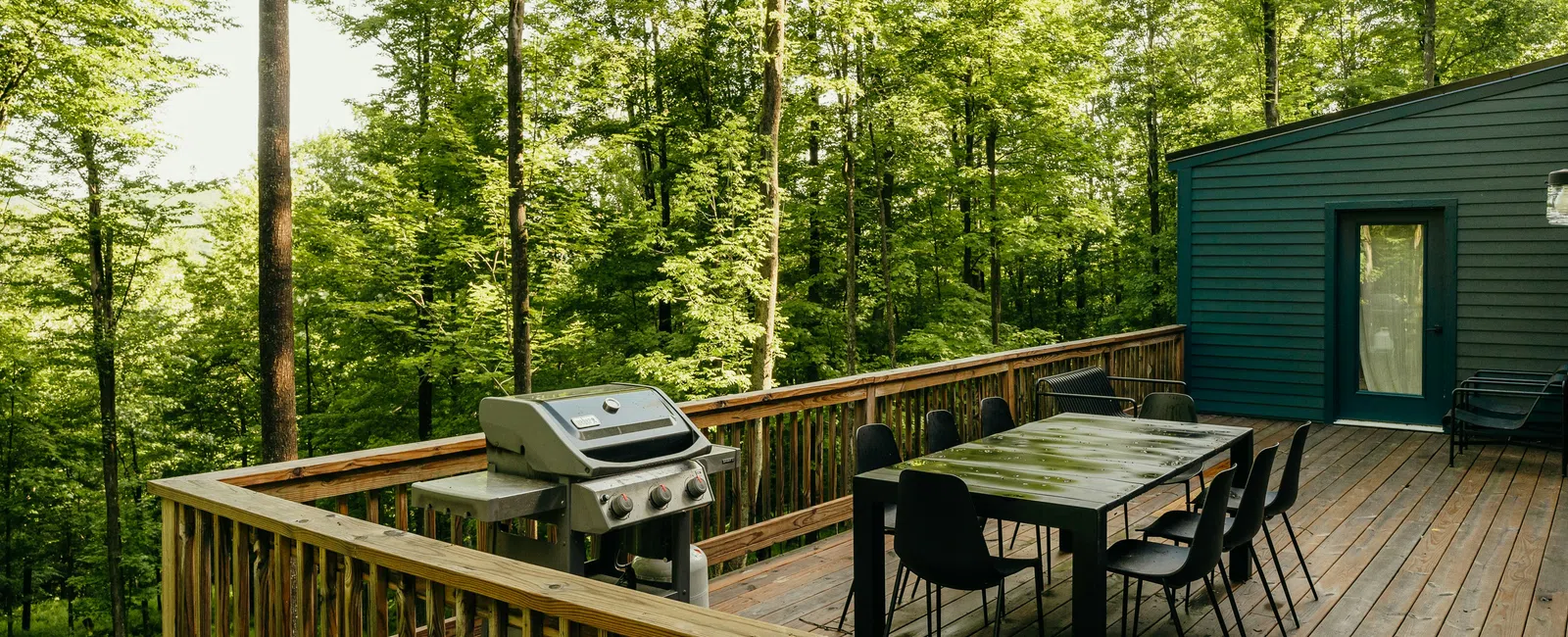 Wooden deck with dining table, chairs, and grill surrounded by dense green forest on a sunny day