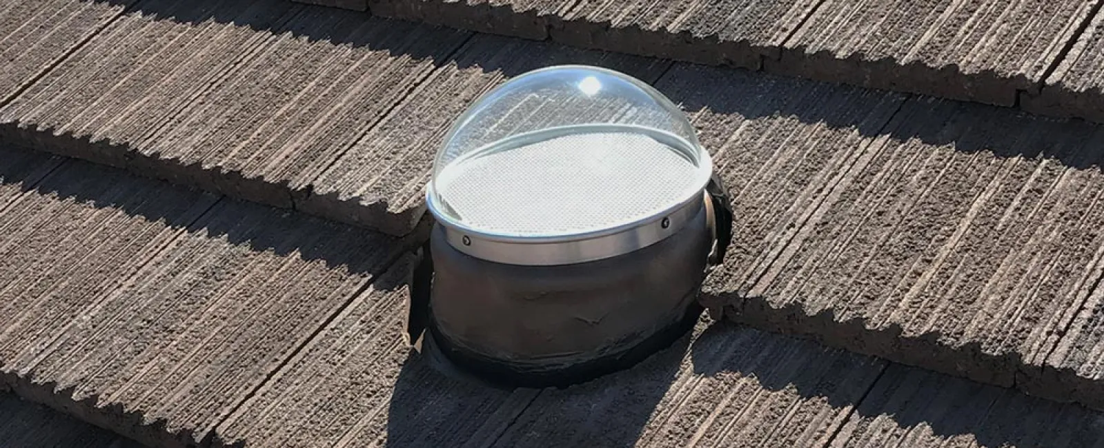 Clear dome skylight installed on textured brown roof tiles casting shadows in sunlight