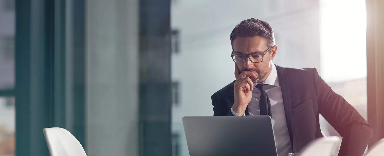 Businessman in suit working on laptop in modern office with large windows and natural light