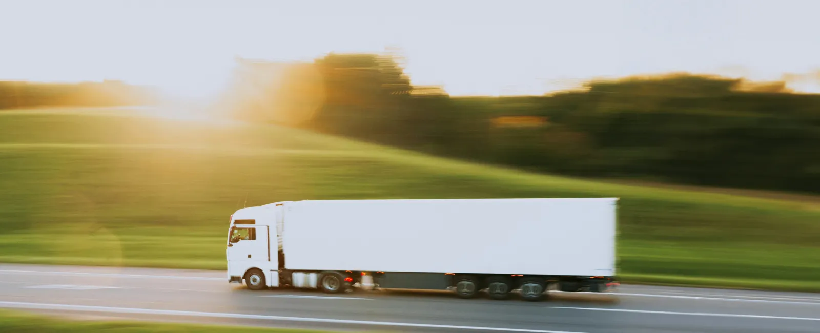 White semi-truck driving fast on a highway through green fields at sunset with motion blur effect.