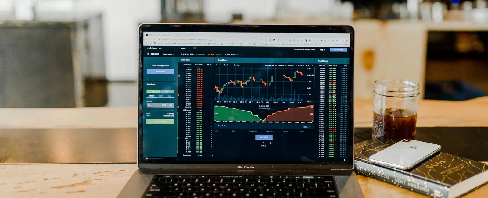 Laptop on table displaying stock market charts and data with a smartphone and drink nearby in a cozy workspace.