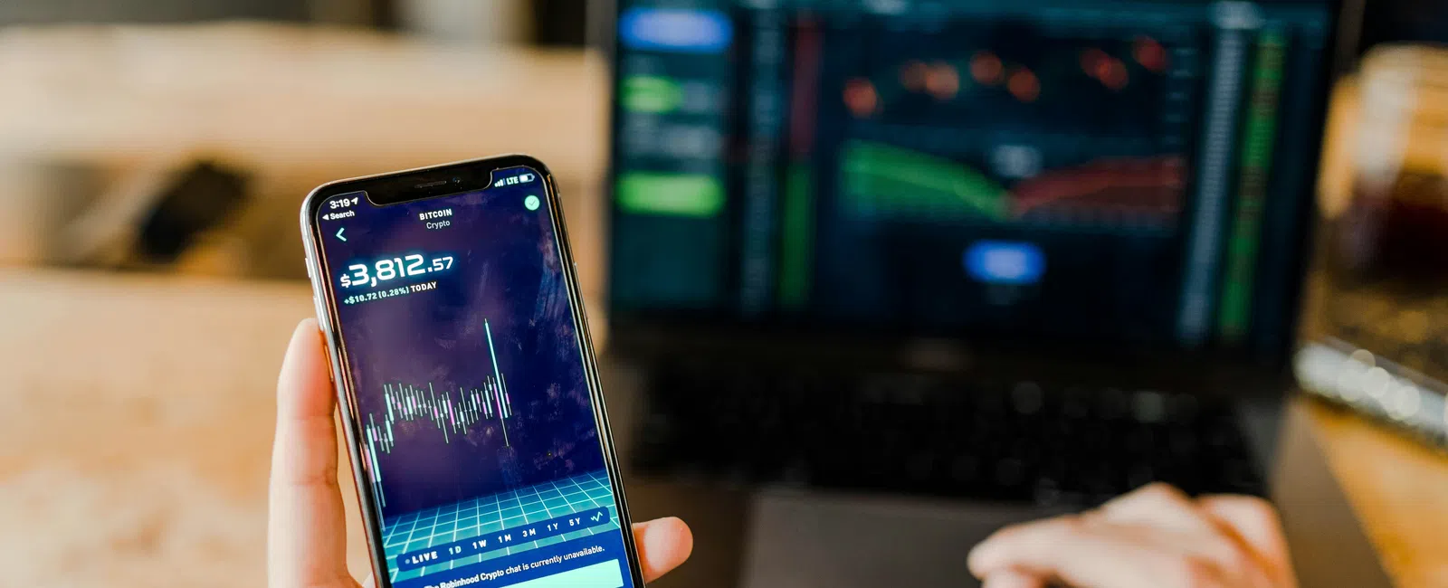 Person holding smartphone showing cryptocurrency trading app with price and graph, laptop with trading data in background.