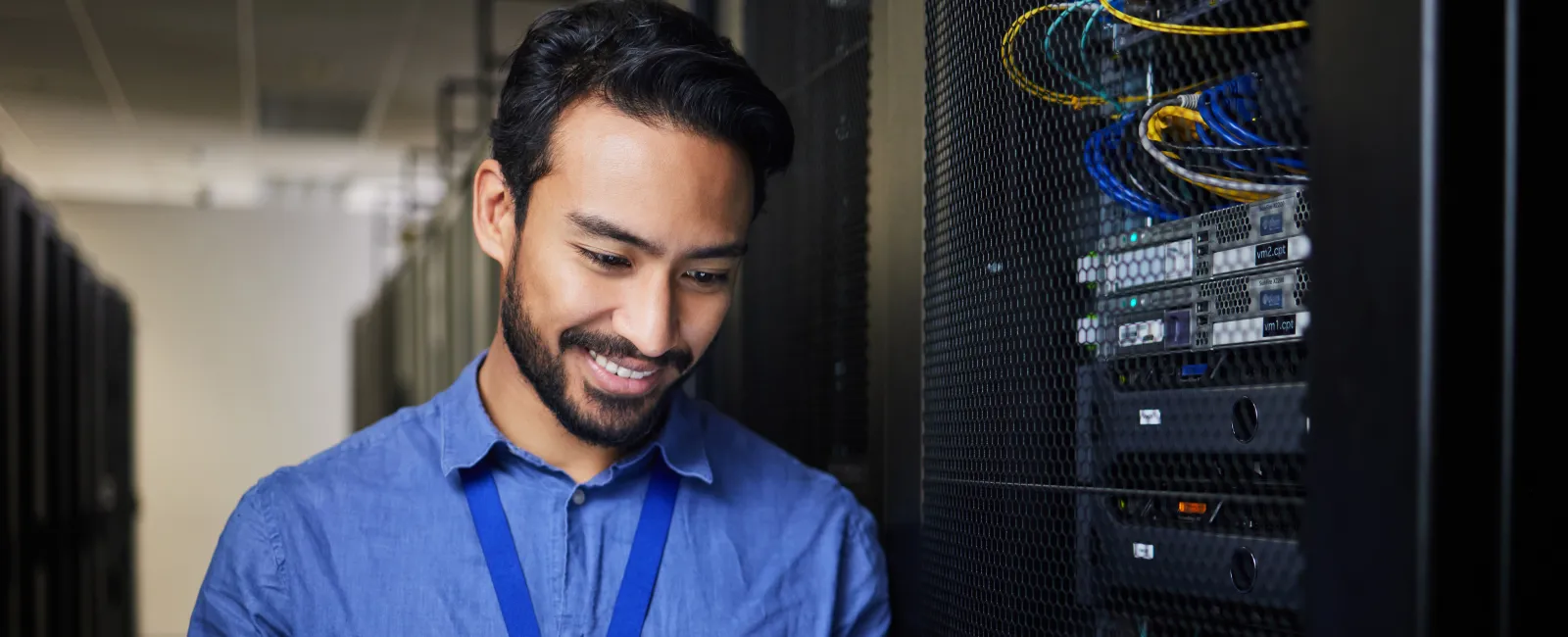 IT technician smiling while using a tablet in a server room with network cables and equipment.