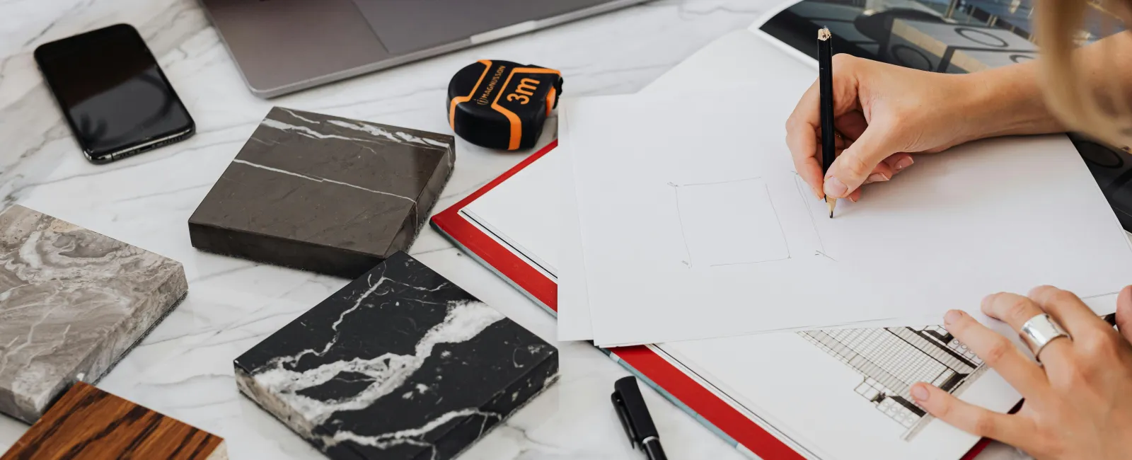Designer sketching building plans surrounded by marble and wood samples on a white table.