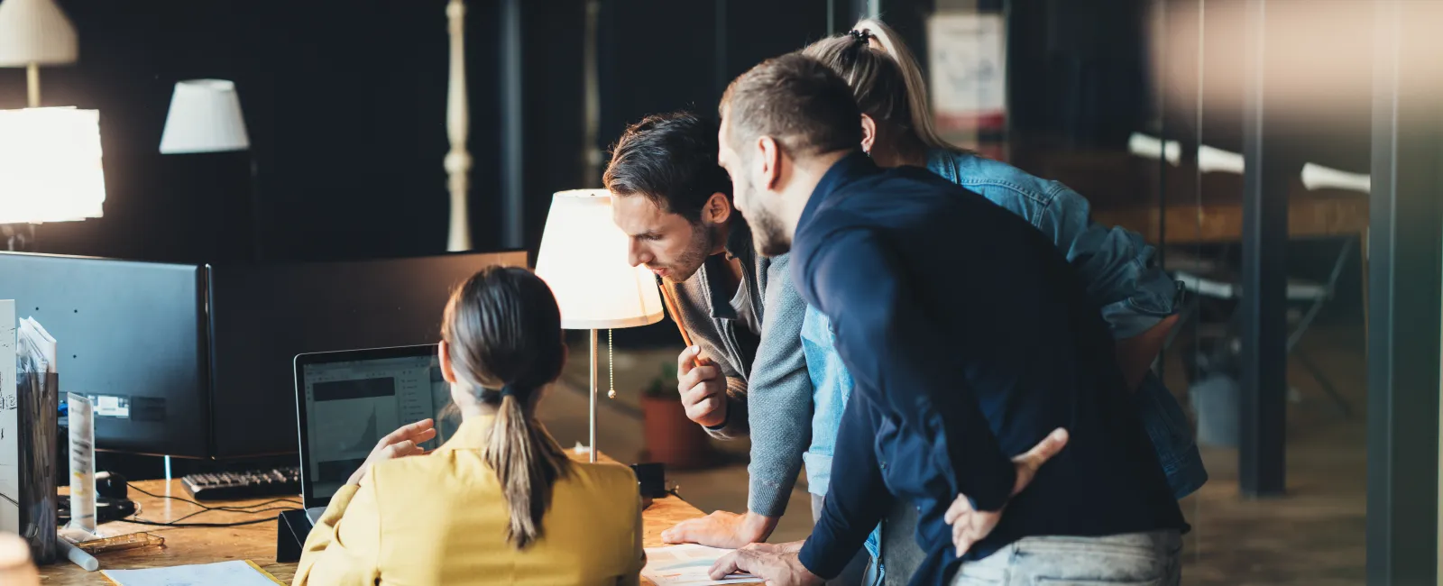Four colleagues collaborating around a laptop in a modern office with warm lighting and wooden desks.