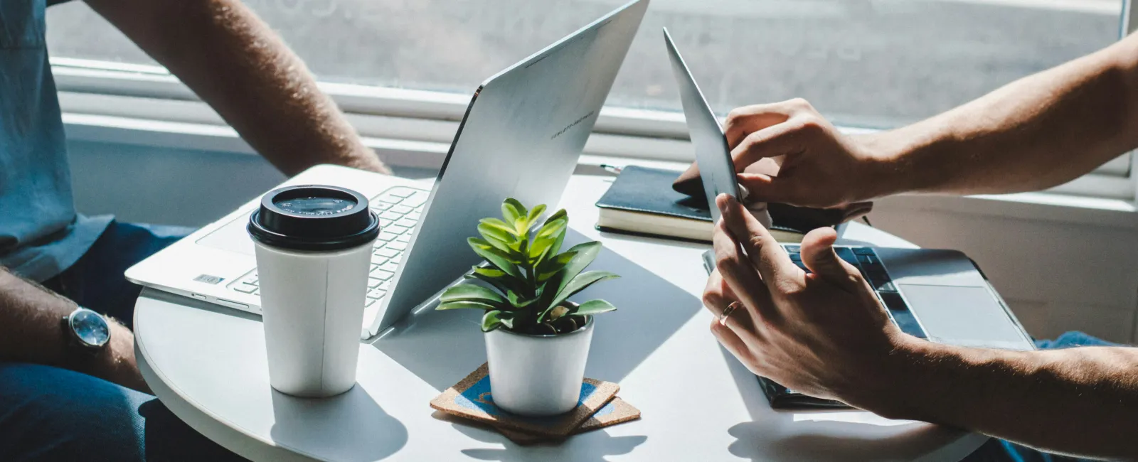 Two people working at a round table with laptop, tablet, coffee cup, and small plant near a window with sunlight.