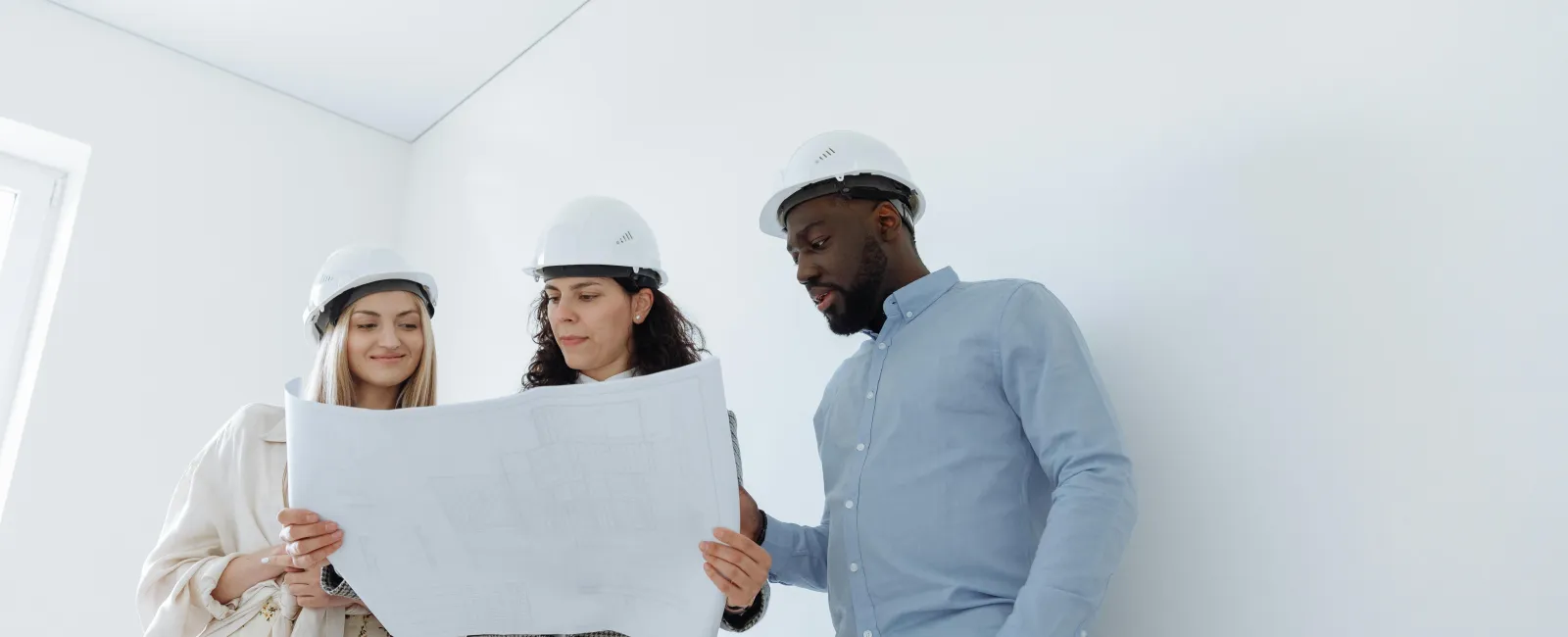 Three architects wearing helmets reviewing building blueprints in a bright, empty room with white walls.