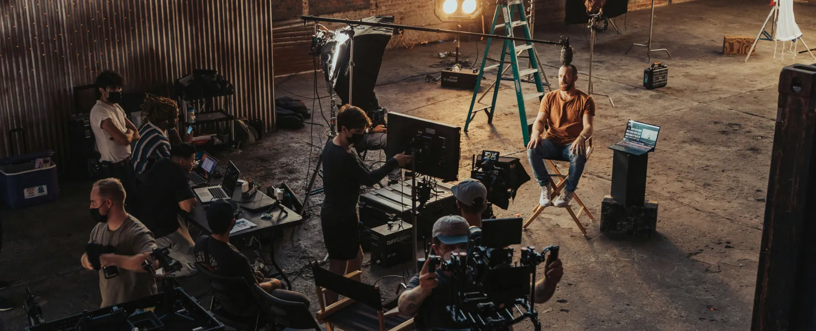 Film crew filming a man sitting on a chair in a large industrial studio with professional lighting and camera equipment