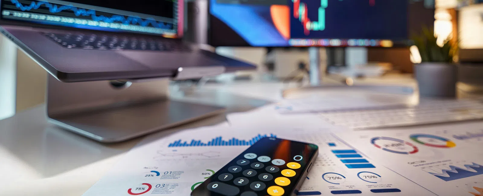 Smartphone with calculator app on desk surrounded by financial charts and dual monitors displaying stock market graphs