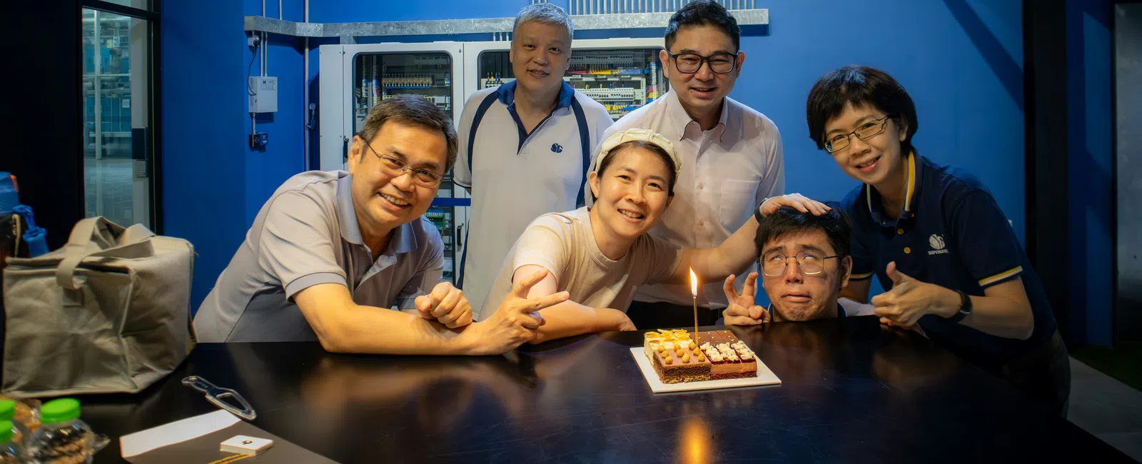 Group of six people smiling around a birthday cake with a lit candle in an indoor setting with blue walls.