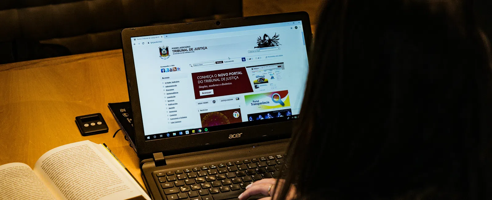 Person using Acer laptop displaying Tribunal de Justiça website with an open book on a wooden table.
