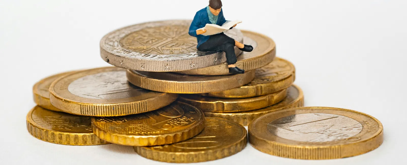 Miniature figure sitting and reading on a stack of gold and silver coins symbolizing financial knowledge.