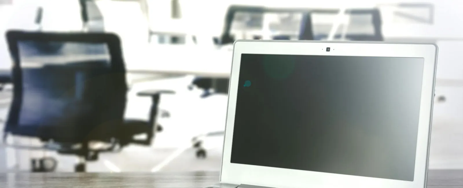 Silver laptop with black keyboard on wooden desk in bright modern office with blurred chairs in background