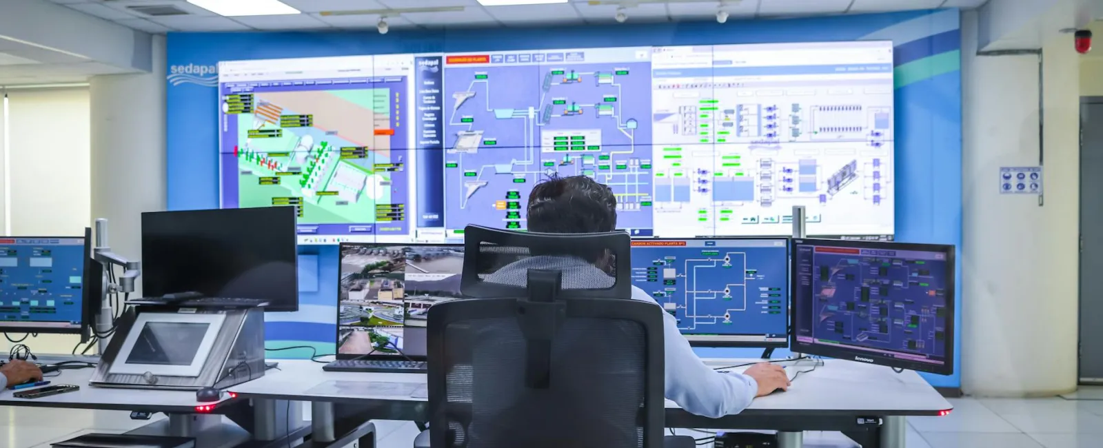 Operator monitoring multiple computer screens displaying control system diagrams and data in a modern control room.